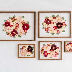 May include: Five rectangular wooden frames containing pressed flower arrangements. Each frame displays a collection of dried flowers in shades of deep red, light pink, and white, accented with green leaves, set against a white background.