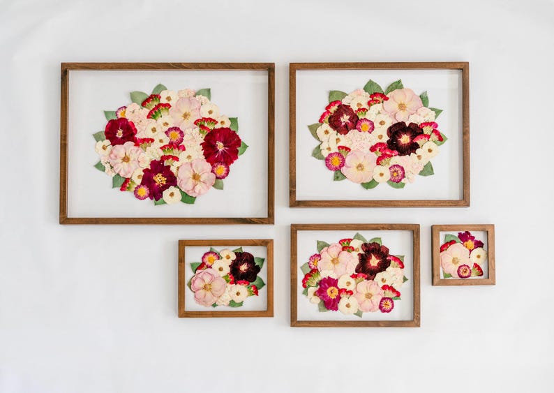 May include: Five rectangular wooden frames containing pressed flower arrangements. Each frame displays a collection of dried flowers in shades of deep red, light pink, and white, accented with green leaves, set against a white background.