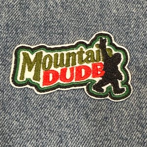 May include: An embroidered patch featuring the text Mountain in olive green and DUDE in bright red. To the right of the text is a black silhouette of a creature walking with one arm raised. The entire design is outlined in dark green and white.