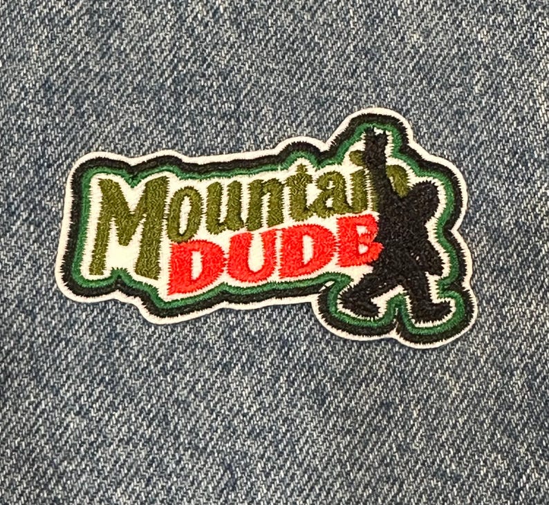 May include: An embroidered patch featuring the text Mountain in olive green and DUDE in bright red. To the right of the text is a black silhouette of a creature walking with one arm raised. The entire design is outlined in dark green and white.