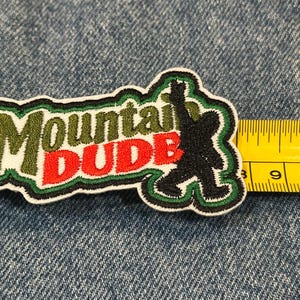 May include: An embroidered patch featuring the text Mountain DUDE in olive green and red thread, accompanied by a black silhouette of a creature. A yellow measuring tape is positioned along the right edge, showing a width of approximately 4 inches.