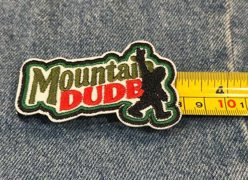 May include: An embroidered patch featuring the text Mountain DUDE in olive green and red thread, accompanied by a black silhouette of a creature. A yellow measuring tape is positioned along the right edge, showing a width of approximately 4 inches.