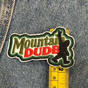 May include: An embroidered patch featuring the text Mountain DUDE in green and red thread with a black silhouette of a creature. A yellow measuring tape shows the patch is approximately 3.5 inches wide and 2 inches tall at its widest points.