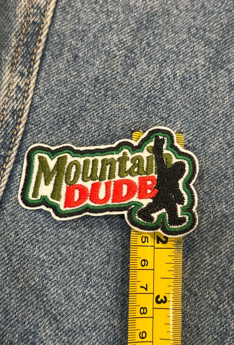 May include: An embroidered patch featuring the text Mountain DUDE in green and red thread with a black silhouette of a creature. A yellow measuring tape shows the patch is approximately 3.5 inches wide and 2 inches tall at its widest points.