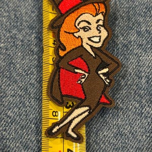 May include: An embroidered patch of a cartoon witch with orange hair, wearing a brown dress and hat with red accents, positioned against a yellow measuring tape showing a height of approximately 10 cm from the top of the hat to the feet.