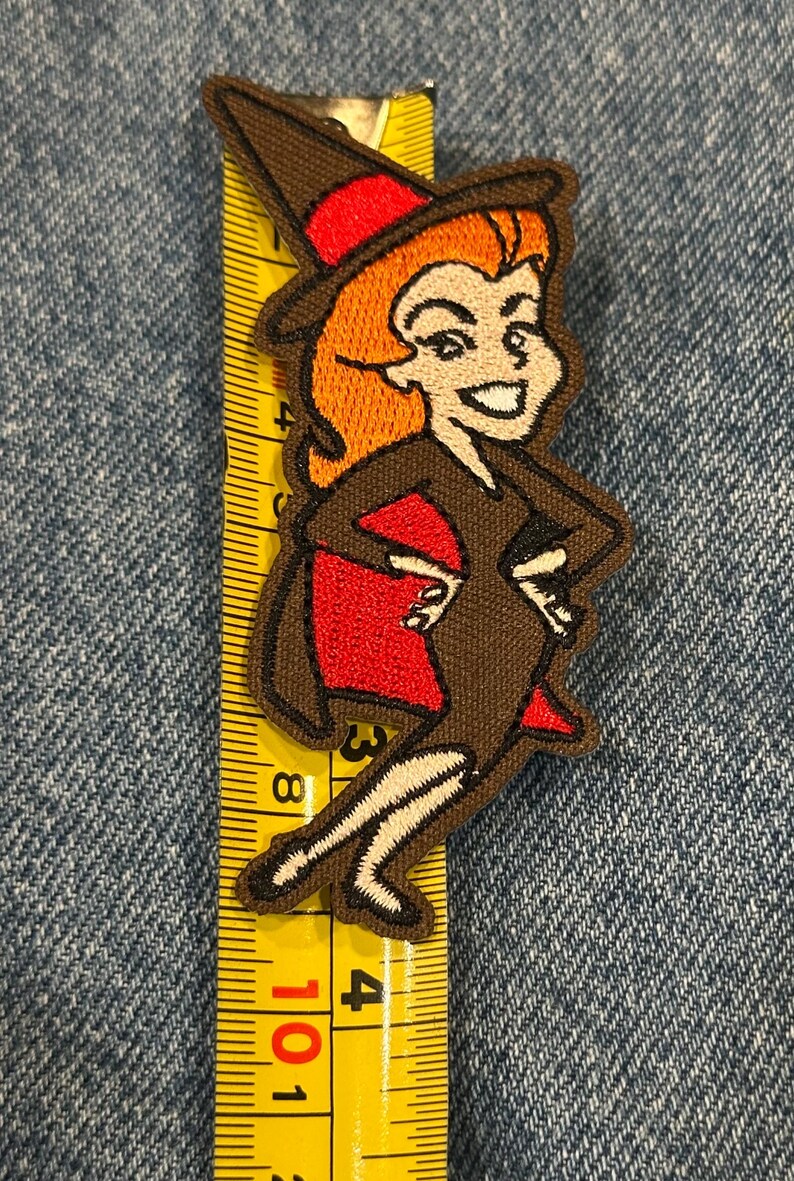 May include: An embroidered patch of a cartoon witch with orange hair, wearing a brown dress and hat with red accents, positioned against a yellow measuring tape showing a height of approximately 10 cm from the top of the hat to the feet.