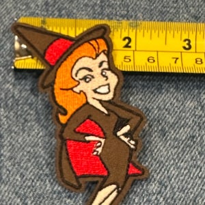 May include: An embroidered patch featuring a cartoon witch with orange hair, a brown dress, and a red cape, positioned next to a yellow measuring tape showing a height of approximately 9 cm from the top of the hat to the feet.