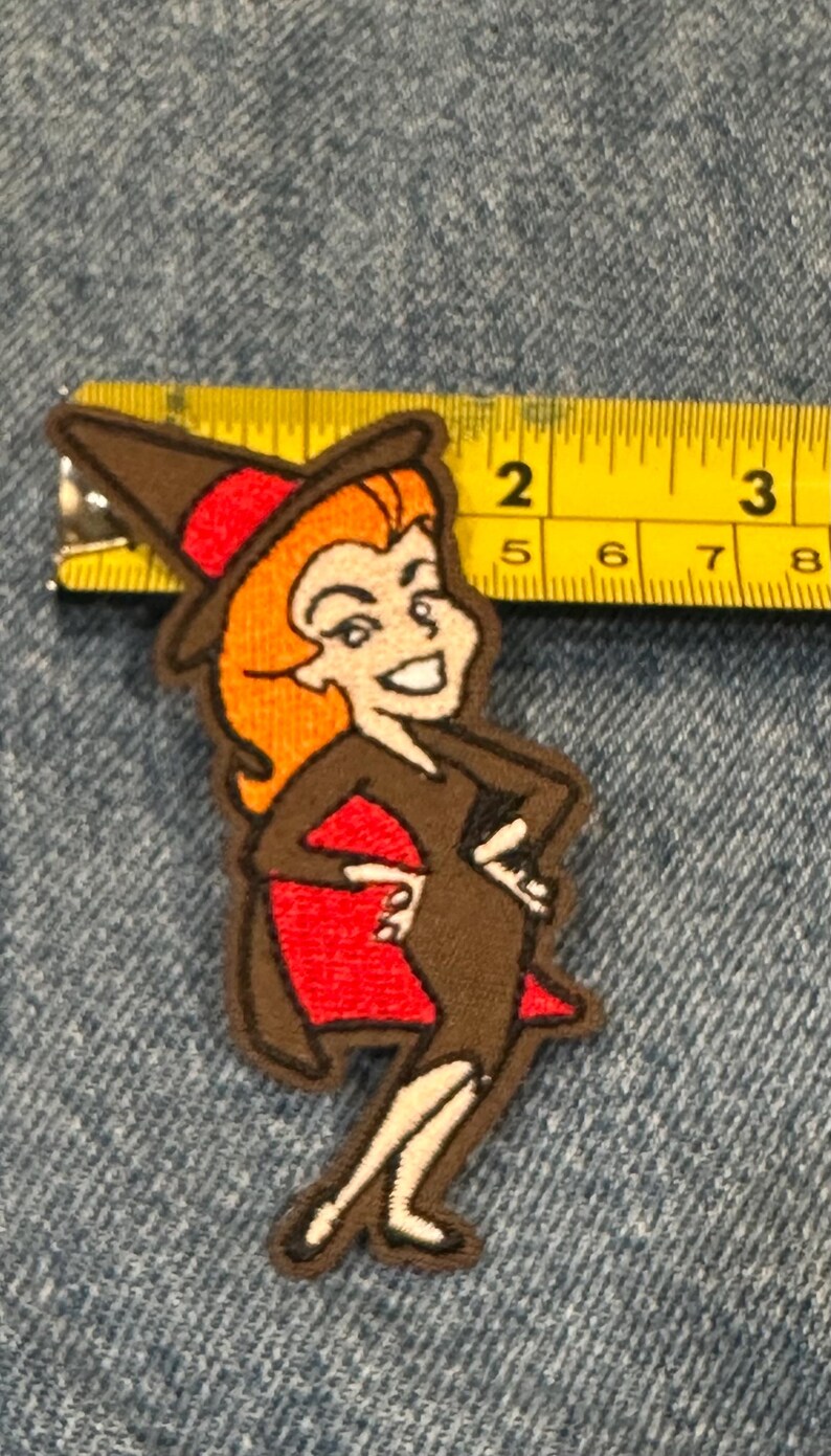 May include: An embroidered patch featuring a cartoon witch with orange hair, a brown dress, and a red cape, positioned next to a yellow measuring tape showing a height of approximately 9 cm from the top of the hat to the feet.