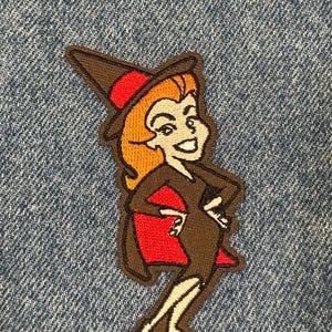 May include: An embroidered patch featuring a cartoon witch with orange hair, wearing a brown pointed hat with a red band, a brown dress, and a red cape. The character is depicted in a standing pose with hands on hips, outlined in dark brown thread.