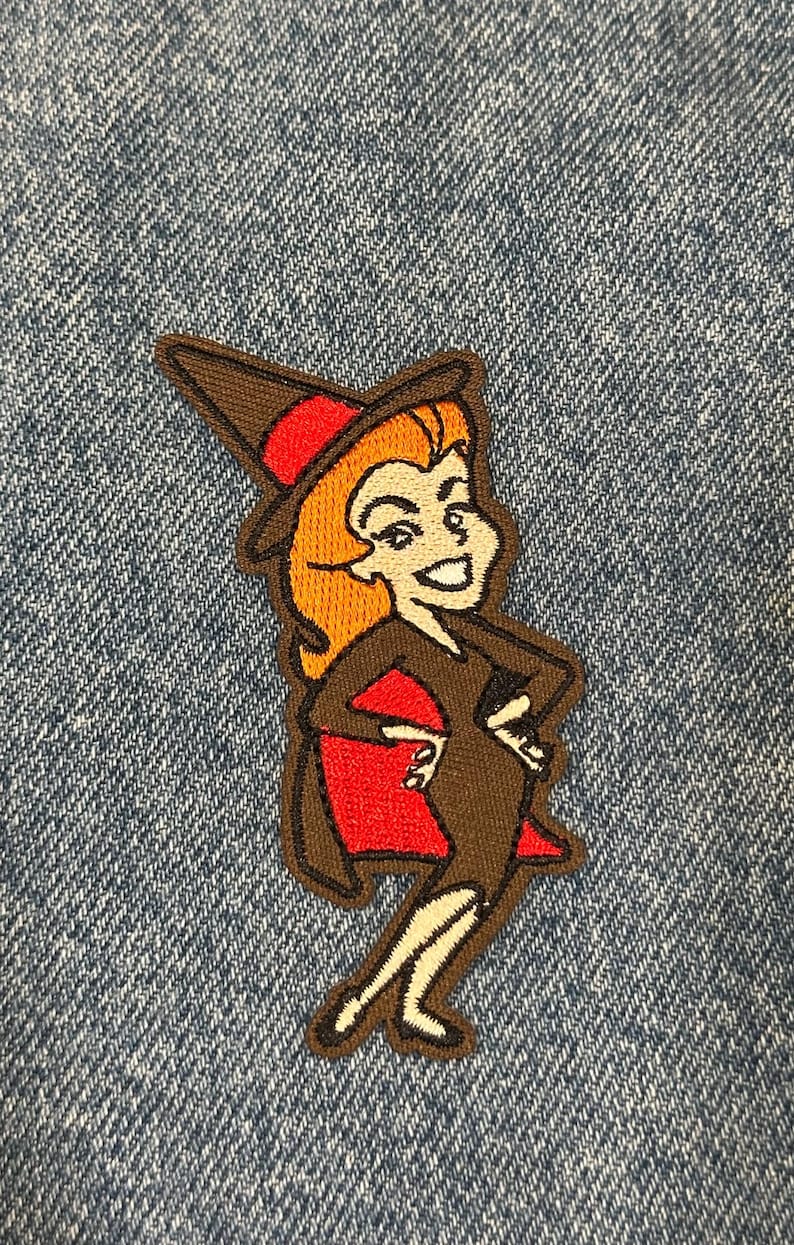 May include: An embroidered patch featuring a cartoon witch with orange hair, wearing a brown pointed hat with a red band, a brown dress, and a red cape. The character is depicted in a standing pose with hands on hips, outlined in dark brown thread.