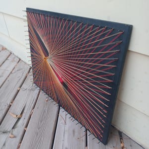May include: A rectangular black board featuring string art created with thin threads in shades of red, orange, and cream. The threads are wrapped around small metal nails driven into the edges of the board, forming a geometric pattern.