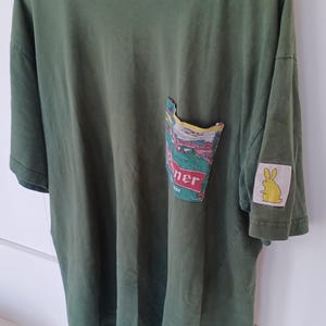 May include: Olive green t-shirt with a pocket featuring a colourful graphic and the word "ner" in red. A yellow bunny patch is on the sleeve. The shirt is hanging on a white hanger.