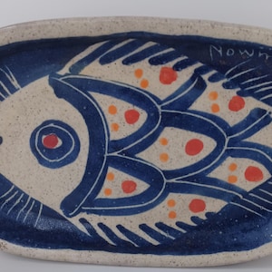 Peut inclure: Plat de service ovale en céramique avec un motif de poisson. Le poisson est représenté en bleu et orange sur un fond beige, avec une bordure bleu foncé. La signature de l'artiste, "Nowmski", est visible sur le bord.