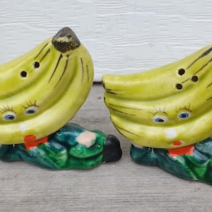 May include: A pair of ceramic salt and pepper shakers shaped like yellow bananas with painted blue eyes, eyelashes, and red mouths. The banana figures are depicted lying down on green bases with black shoes and small pink hands visible on the sides.
