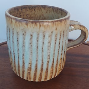 May include: A handmade ceramic mug with a textured, vertical striped design. The mug features a light blue and tan colour scheme with brown accents. The handle is curved and integrated into the mug's design. This unique mug is perfect for coffee or tea.