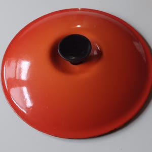 May include: A round, vibrant orange enameled cast iron lid featuring a smooth, glossy finish and a centered, circular black knob. The lid has a slightly domed shape with a clean, polished edge, designed to fit securely onto a matching cookware piece.