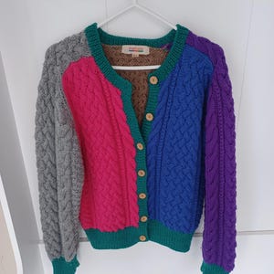May include: A colorful, cable-knit cardigan with a patchwork design. The sweater features panels of gray, pink, blue, and purple, with green trim and wooden buttons. The label reads "PORTLAND".