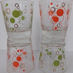 May include: Two clear drinking glasses with a retro design. Each glass features a pattern of orange, green, and brown circles. The glasses are stacked, displaying the design on both sides.