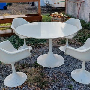 May include: A white, mid-century modern dining set. The set comprises a round table and four matching chairs. The table and chairs have a pedestal base. The chairs have a curved, bucket-style seat.