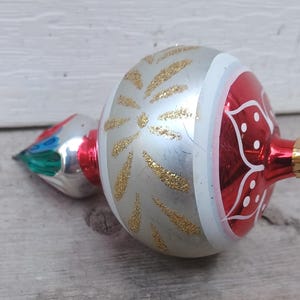 May include: A shiny, teardrop-shaped Christmas ornament featuring a silver centre band decorated with gold glitter starburst patterns. The ornament has a glossy red section with white floral line art and a gold-toned metal cap for hanging.