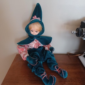 May include: A vintage doll dressed in a teal velvet elf costume with a matching pointed hat and cape. The doll's outfit includes a pink and white floral patterned shirt and matching teal velvet pants. The doll is seated on a wooden surface.