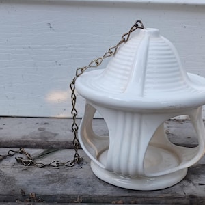 May include: A white ceramic hanging bird feeder with a domed, ribbed roof and a circular base featuring four arched openings. The structure is suspended by a thin, metallic chain attached to the top centre, providing a clear view of the interior.