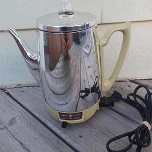 Peut inclure: Une cafetière électrique à percolateur General Electric vintage avec un corps en chrome poli, une poignée en plastique couleur crème et une base assortie. La base comporte un cadran de contrôle marqué MILD, MED et STRONG. Cordon noir inclus.