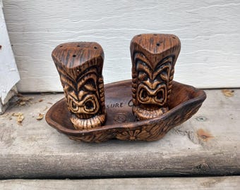 Könnte beinhalten: Ein Paar Salz- und Pfefferstreuer aus brauner Keramik in Form von Tiki-Figuren, die in einer passenden, bootförmigen Keramikschale stehen. Die Schale hat ein florales Reliefmuster an der Außenseite und das Wort "SURE" auf dem Boden eingeprägt.