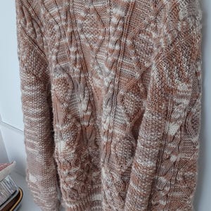 May include: A brown and white cable knit sweater with a round neckline. The sweater is hanging on a white hanger. The knit pattern includes diamond and vertical cable designs.