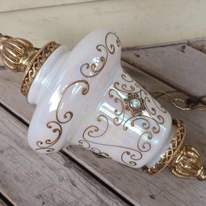 Peut inclure: Une lampe suspendue dotée d'un abat-jour en verre blanc irisé orné de motifs de volutes en filigrane dorés. Le luminaire comprend des embouts en métal doré ouvragé aux deux extrémités et un accent floral central avec un cristal clair.