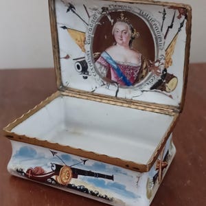 May include: A rectangular, open trinket box with a gold-colored trim. The lid features a portrait of a woman in a crown, surrounded by text and military imagery. The box's sides are decorated with cannons and nautical scenes.