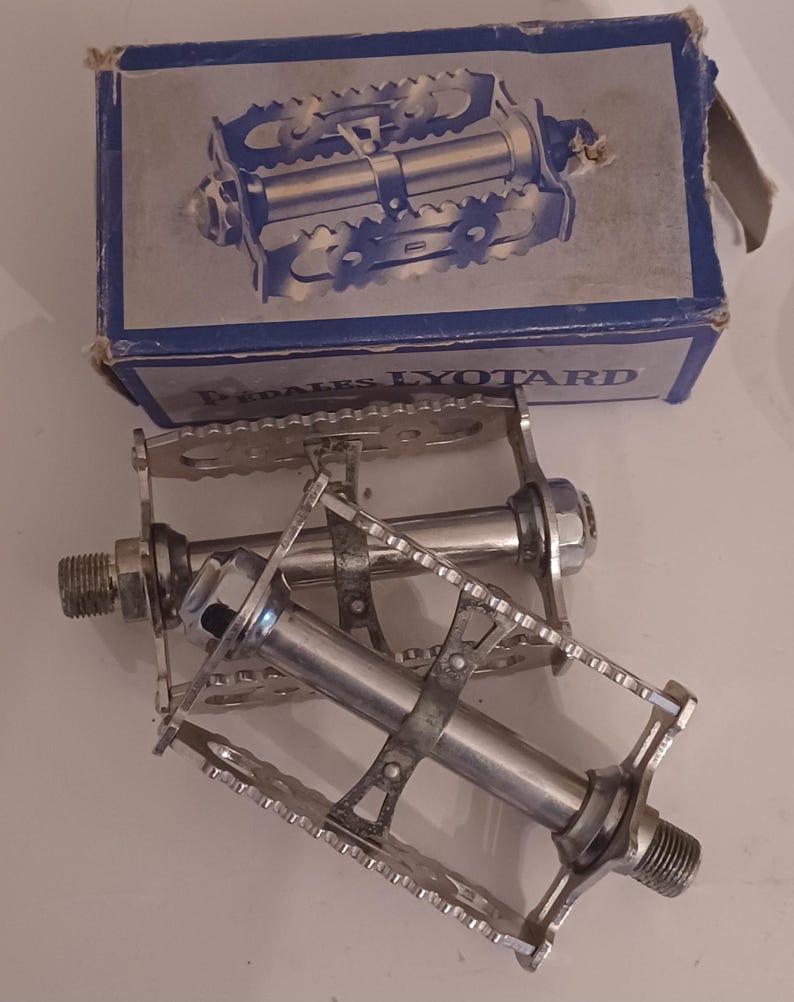 Pu&ograve; includere: Una coppia di pedali per bicicletta argentati con la scritta "PEDALES LYOTARD" sulla scatola.