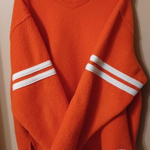 May include: Orange fleece sweatshirt with white stripes on the sleeves and "ROOTS" logo on the front and back.