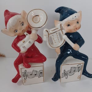 May include: Two ceramic elf salt and pepper shakers. One elf in a red suit plays a white tuba, sitting on a white cube with musical notes and the letter P. The other elf in a blue suit plays a white lyre, sitting on a white cube with musical notes and the letter S.