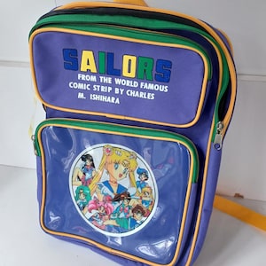 May include: Purple backpack with the word "SAILORS" in blue letters. The backpack features a clear pocket with a cartoon illustration of anime characters. The text "FROM THE WORLD FAMOUS COMIC STRIP BY CHARLES M. ISHIHARA" is printed on the top pocket.