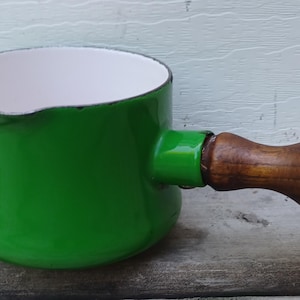May include: A small, round, green enameled metal saucepan with a white interior, a pouring spout on the rim, and a bulbous, polished brown wooden handle attached to the side. The exterior finish is glossy, and the rim shows a thin black edge.