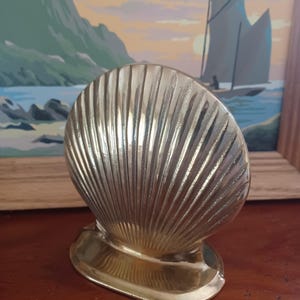May include: A polished brass shell-shaped object with a vertical ribbed texture and a smooth, rounded base. The metallic surface reflects light, highlighting the curved ridges that fan out from the bottom to the top edge of the seashell form.