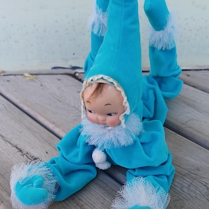 May include: A plush doll dressed in a bright turquoise velvet outfit with a long pointed hat, white lace trim at the collar and cuffs, and white pom-poms. The doll has a painted face with brown hair peeking from under the hood and is posed.