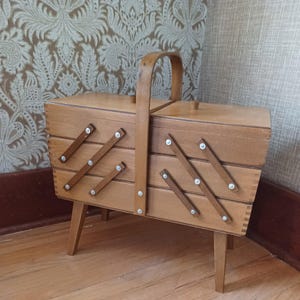 May include: A light brown wooden sewing box featuring a cantilevered accordion-style design with three tiers of storage compartments. The box includes a central arched handle, two top lids with small knobs, and four tapered legs for support.