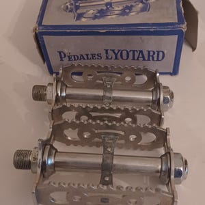 Pu&ograve; includere: Una coppia di pedali per bicicletta argentati con il nome del marchio "P&eacute;dales Lyotard" stampato sulla scatola. I pedali hanno una superficie rigata e un cinturino in metallo.