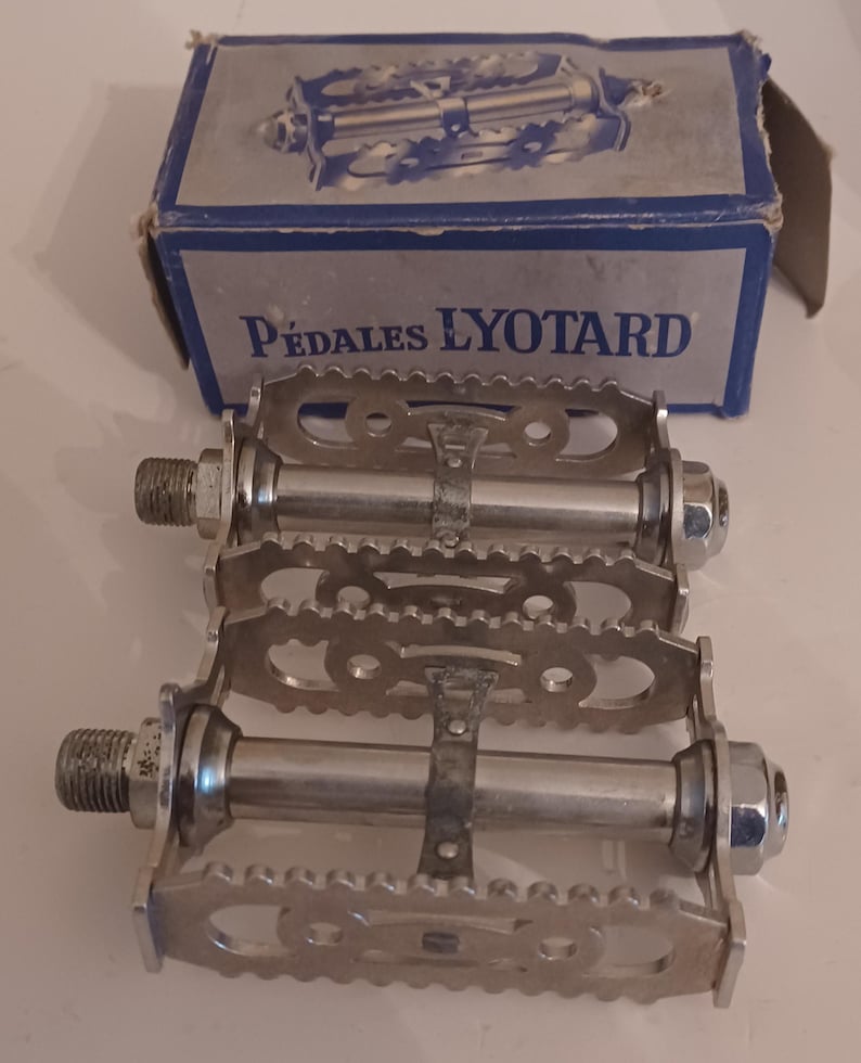 Pu&ograve; includere: Una coppia di pedali per bicicletta argentati con il nome del marchio "P&eacute;dales Lyotard" stampato sulla scatola. I pedali hanno una superficie rigata e un cinturino in metallo.