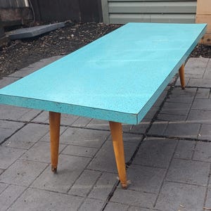 May include: A rectangular turquoise table with speckled surface detail. The table has four tapered, light-brown wooden legs. The table is approximately 152 cm long and 76 cm wide.