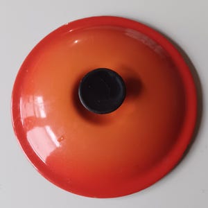May include: A round, enameled cast iron pot lid featuring a vibrant orange gradient finish that transitions from a deep red-orange at the outer rim to a lighter, bright orange toward the center. It includes a solid black, circular knob handle.