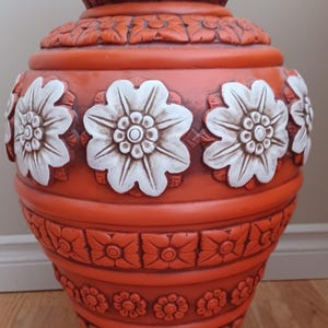 May include: A large, orange ceramic vase with white floral designs. The vase has a wide base and a narrow neck, and is decorated with intricate carvings.