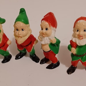 Peut inclure: Quatre figurines de gnomes vintage. Chaque gnome est vêtu d'un manteau rouge, d'un pantalon vert et de chaussures noires. Les gnomes ont des barbes blanches et portent un chapeau vert ou rouge. Un gnome tient une brindille verte.