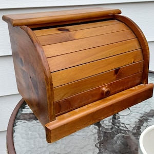 Peut inclure: Une boîte à pain en bois brun moyen dotée d'une porte coulissante à lattes incurvées avec un petit bouton circulaire. Le récipient de rangement présente une base rectangulaire et une surface supérieure plate, avec des veines de bois naturelles.