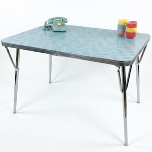 May include: A rectangular folding table with a light blue patterned laminate top and chrome metal legs. A vintage-style teal rotary telephone sits on the left side, and a stack of four small, colorful ceramic cups sits on the right side.