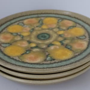 May include: A stack of four ceramic plates. Each plate has a beige rim and a central design featuring yellow, orange, and green floral patterns. A teal-colored band encircles the central design.