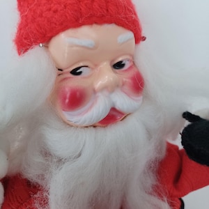 May include: A vintage Santa Claus doll with a red knit hat and suit. The doll has a painted face with rosy cheeks, white eyebrows, and a long white beard. Black gloves and a black belt complete the outfit. A classic Christmas decoration.