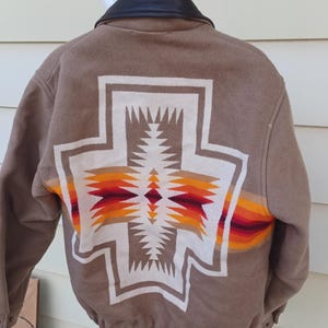 May include: A tan jacket with a black leather collar and a large, centered geometric pattern on the back. The design features a white cross shape filled with orange, yellow, and deep red horizontal stripes, creating a bold, symmetrical visual.